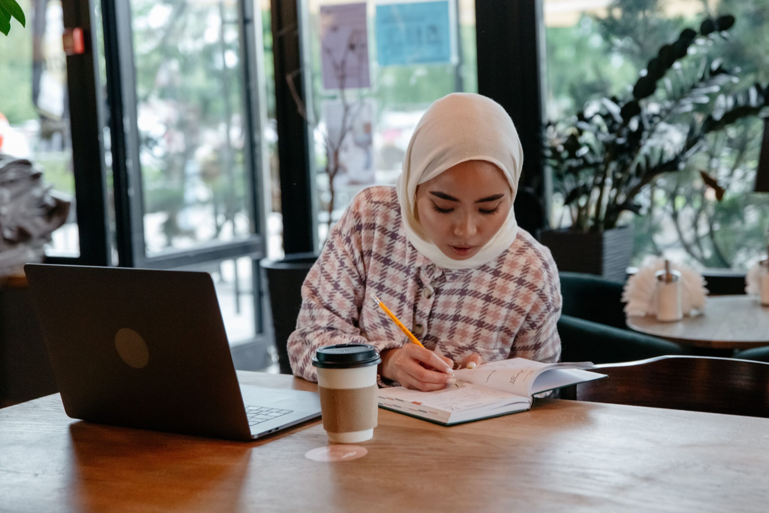 A young Asian woman in a hijab studies at a coffee shop, with a laptop and notebook.